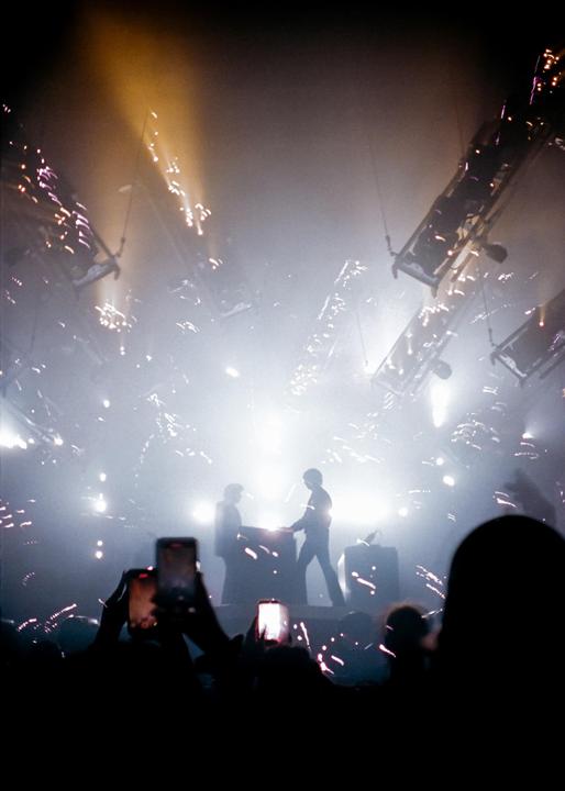 Two men are performing on a stage with bright lights and pyrotechnics while an audience records them with their phones.