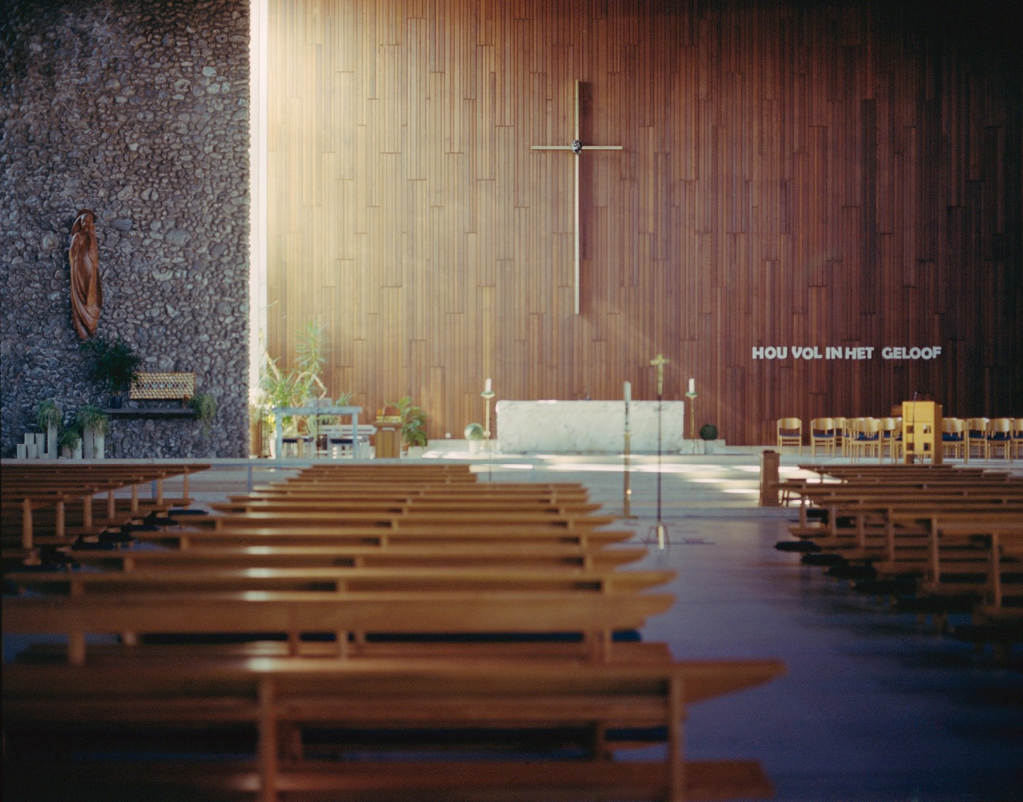 Sunlight streams into a church with wooden pews, a stone altar, and a large cross on a wooden wall.