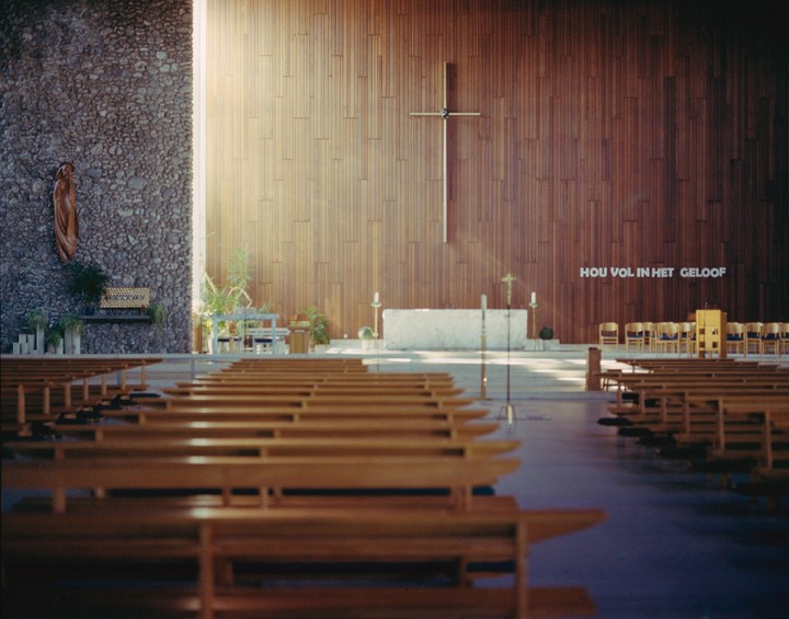 Sunlight streams into a church with wooden pews, a stone altar, and a large cross on a wooden wall.