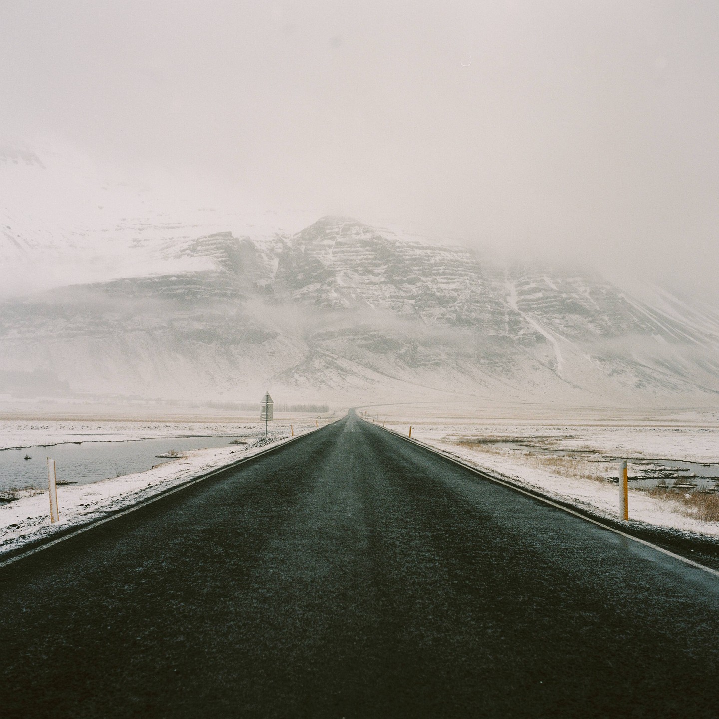 A long, empty road stretches towards a snow-covered mountain range under a hazy sky.