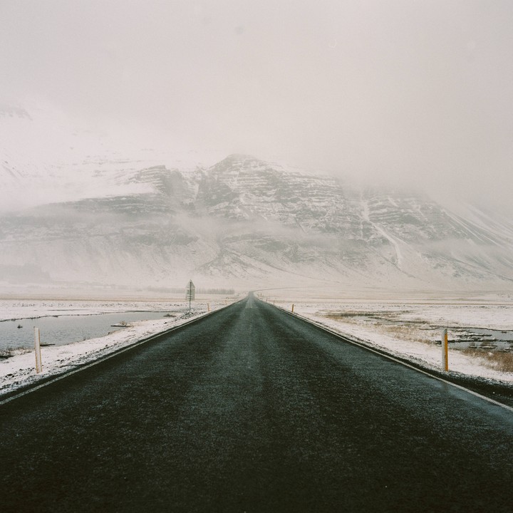 A long, empty road stretches towards a snow-covered mountain range under a hazy sky.
