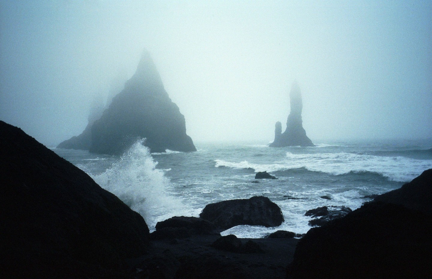Waves crash against dark rocks on a foggy beach with two large sea stacks in the distance.