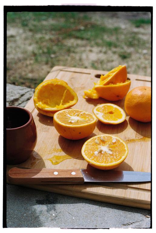 A wooden cutting board with oranges, orange peels, and a knife sits on a stone surface next to a brown cup.