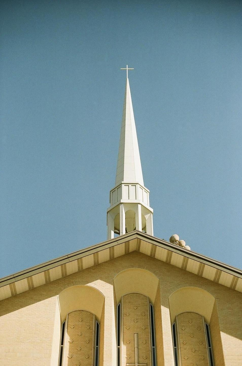 A white steeple with a cross on top rises above a brick building against a clear blue sky.