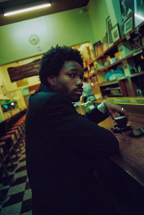 A man with dark, curly hair sits at a bar with a cup of coffee, looking over his shoulder.