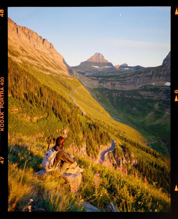 A person with a backpack sits on a rock overlooking a winding road through a mountainous landscape during golden hour.