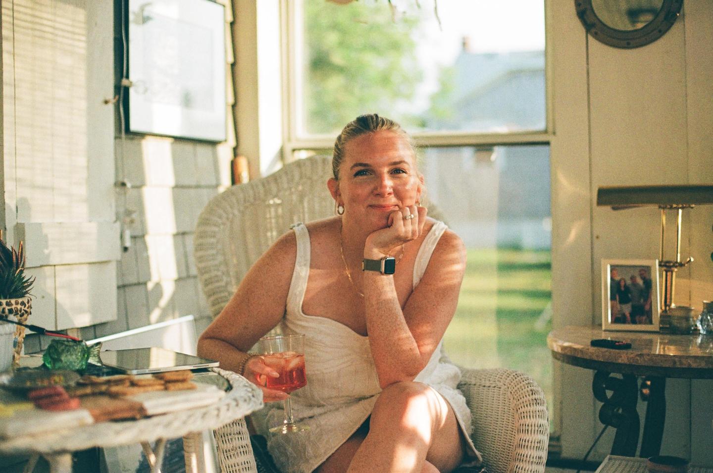 A woman in a white dress sits in a wicker chair, holding a drink and smiling at the camera.