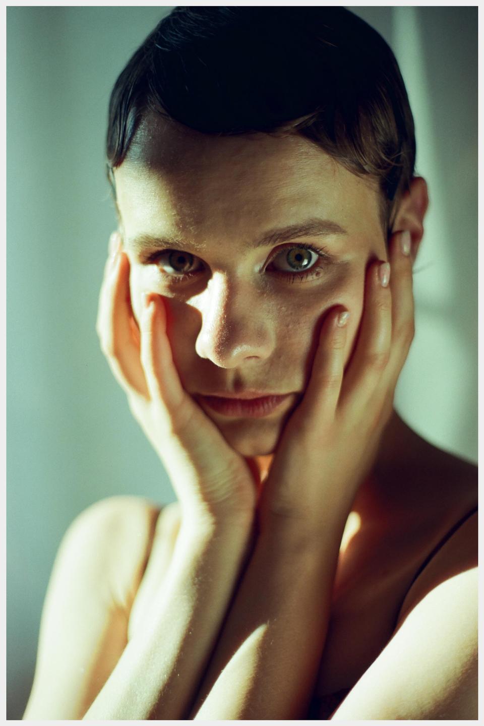 A woman with short hair holds her face in her hands while looking directly at the viewer.