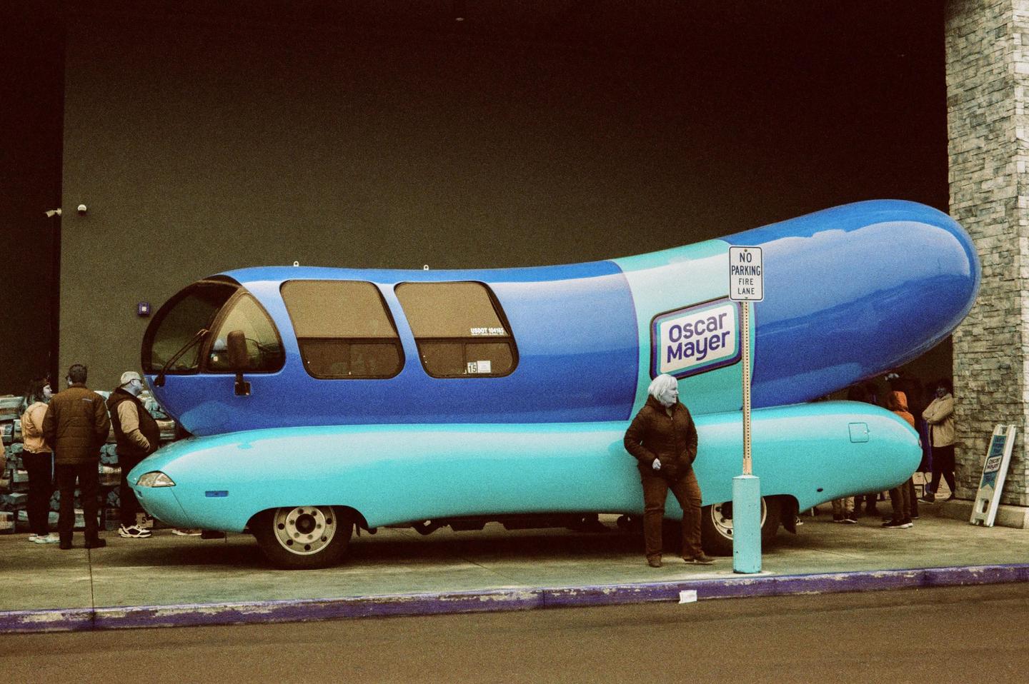 A woman stands next to a large blue and turquoise Oscar Mayer Wienermobile.