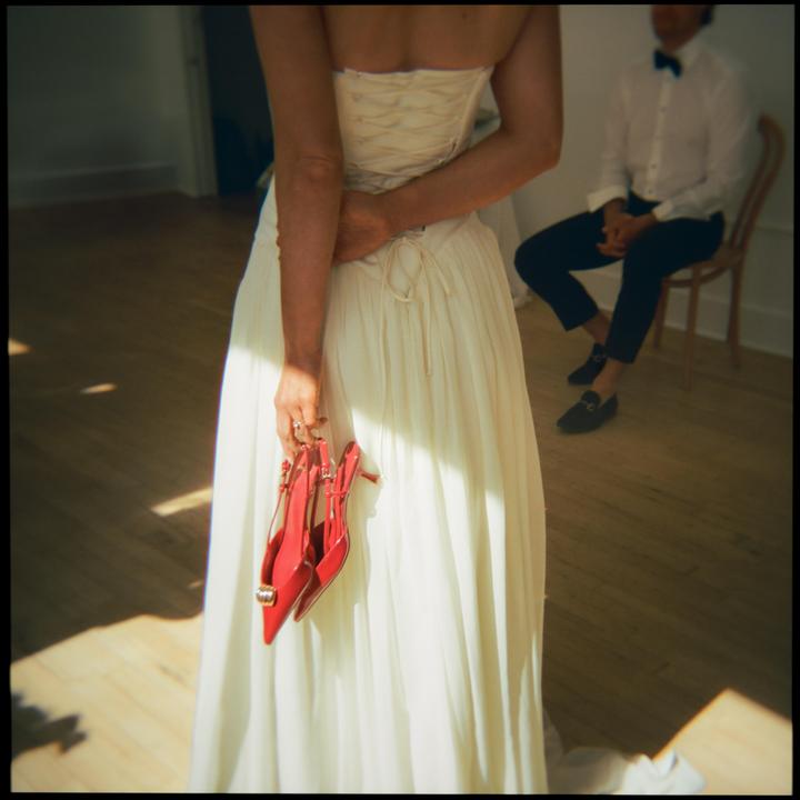 A woman in a white dress holds red high heels behind her back while a man sits in the background.