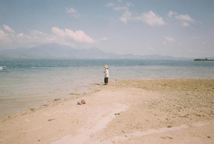 A child wearing a hat stands in shallow water on a sandy beach with mountains in the distance.