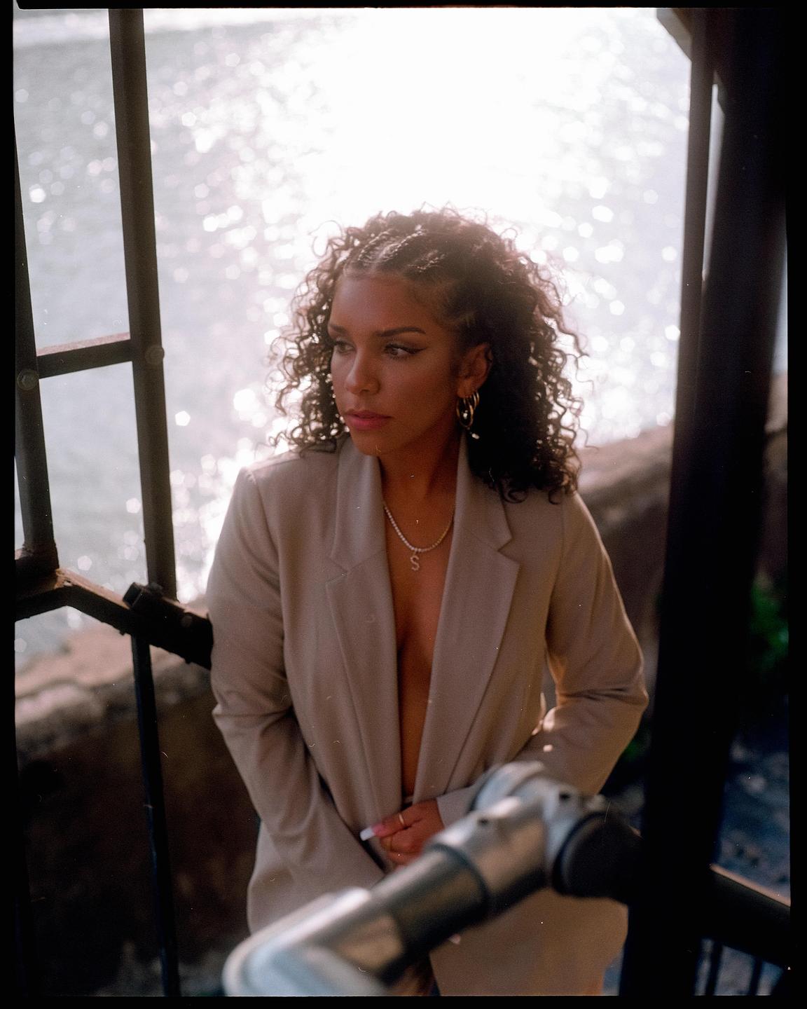 A woman with curly hair and a blazer stands next to a railing with sparkling water in the background.
