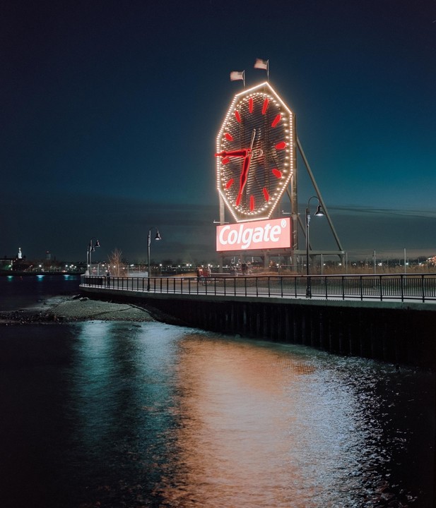 A large illuminated clock and a Colgate sign stand next to a body of water with reflections at night.