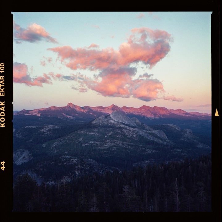 Pink clouds float above a mountain range with a forest in the foreground at dusk.