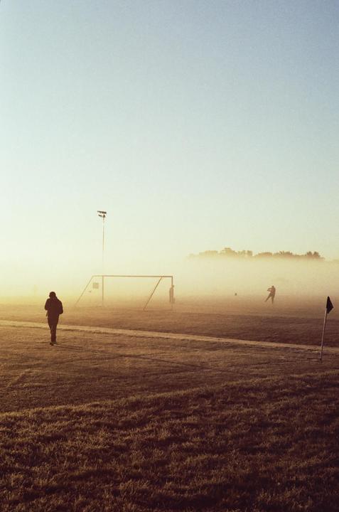 A person walks across a foggy soccer field with a goal and other people in the distance.