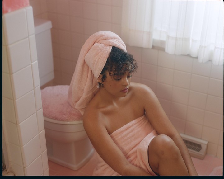 A woman wrapped in a pink towel sits on the floor of a pink tiled bathroom.