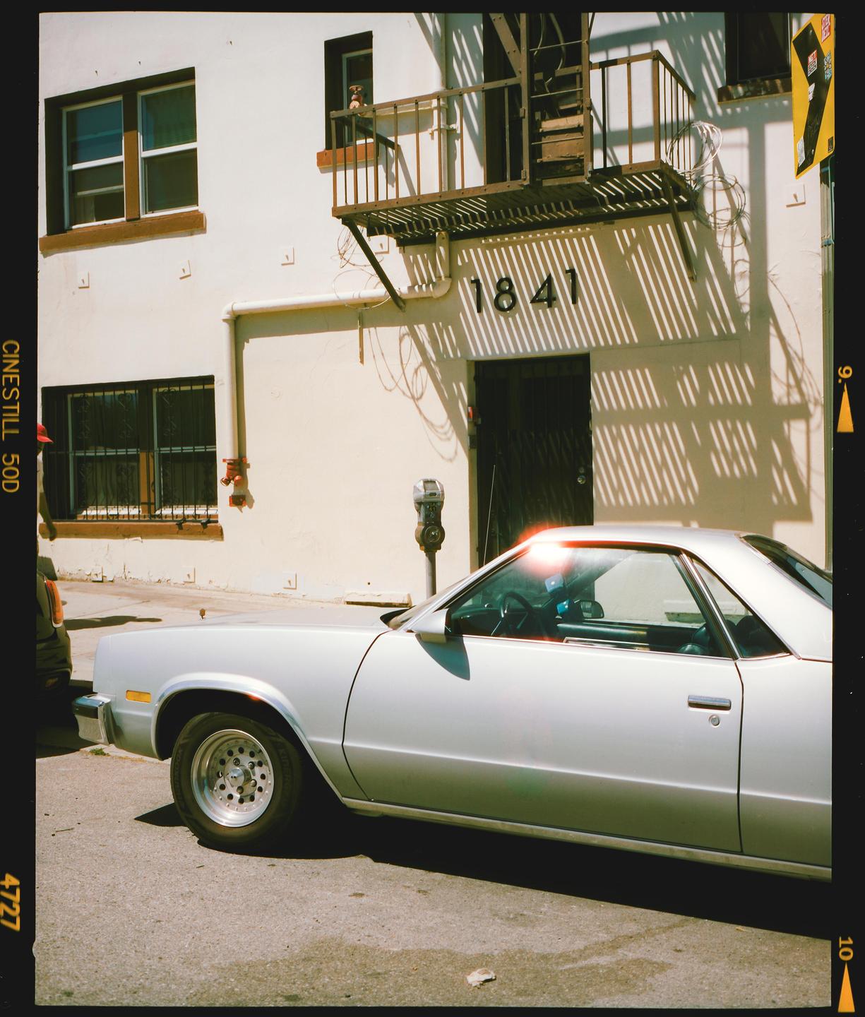 A silver car is parked on a street in front of a building with an external staircase and strong shadows.
