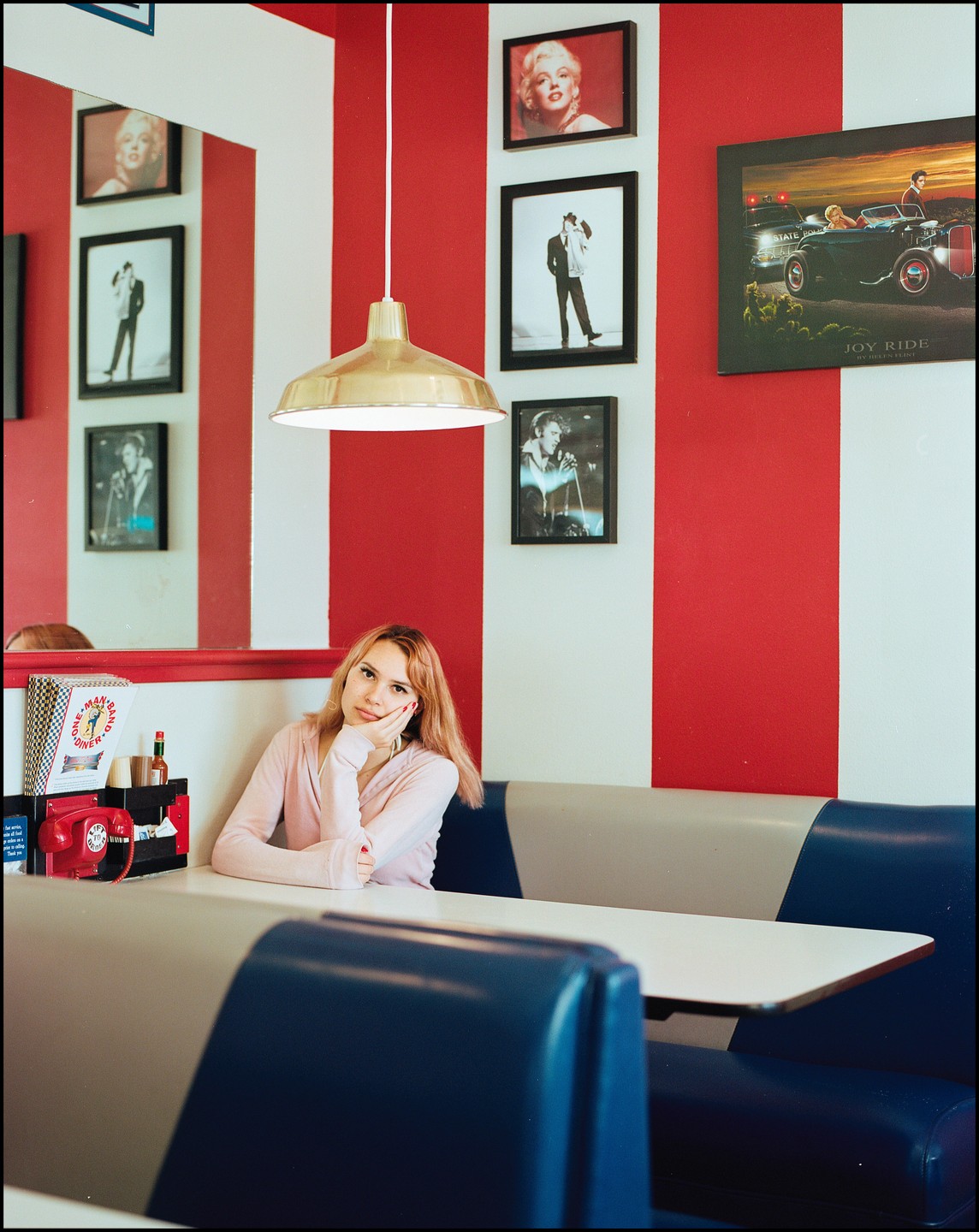 A woman sits in a diner booth with red and white striped walls decorated with framed pictures of celebrities.