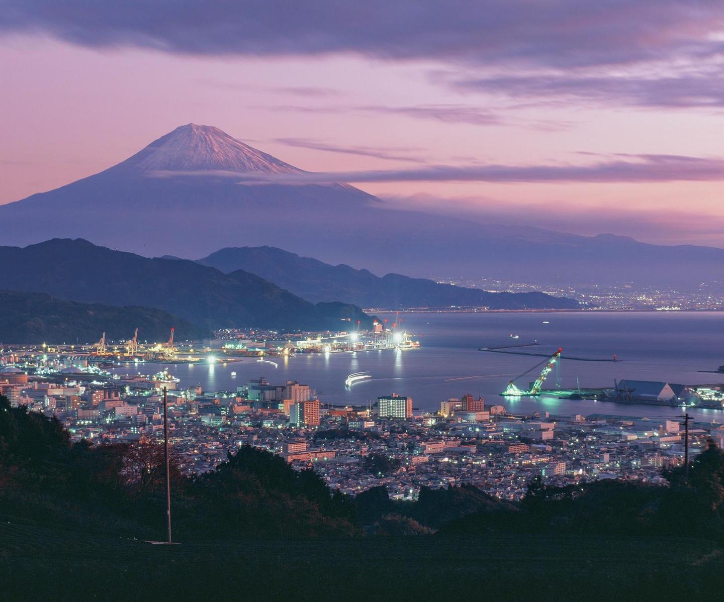 Mount Fuji rises above a city and bay with illuminated buildings and boats at dusk.
