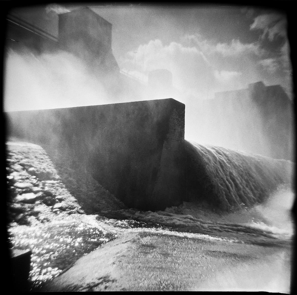 Water flows over a concrete dam, creating mist and spray.