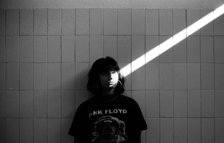 A woman with dark hair and a Pink Floyd t-shirt stands against a tiled wall with a diagonal beam of light illuminating her face.