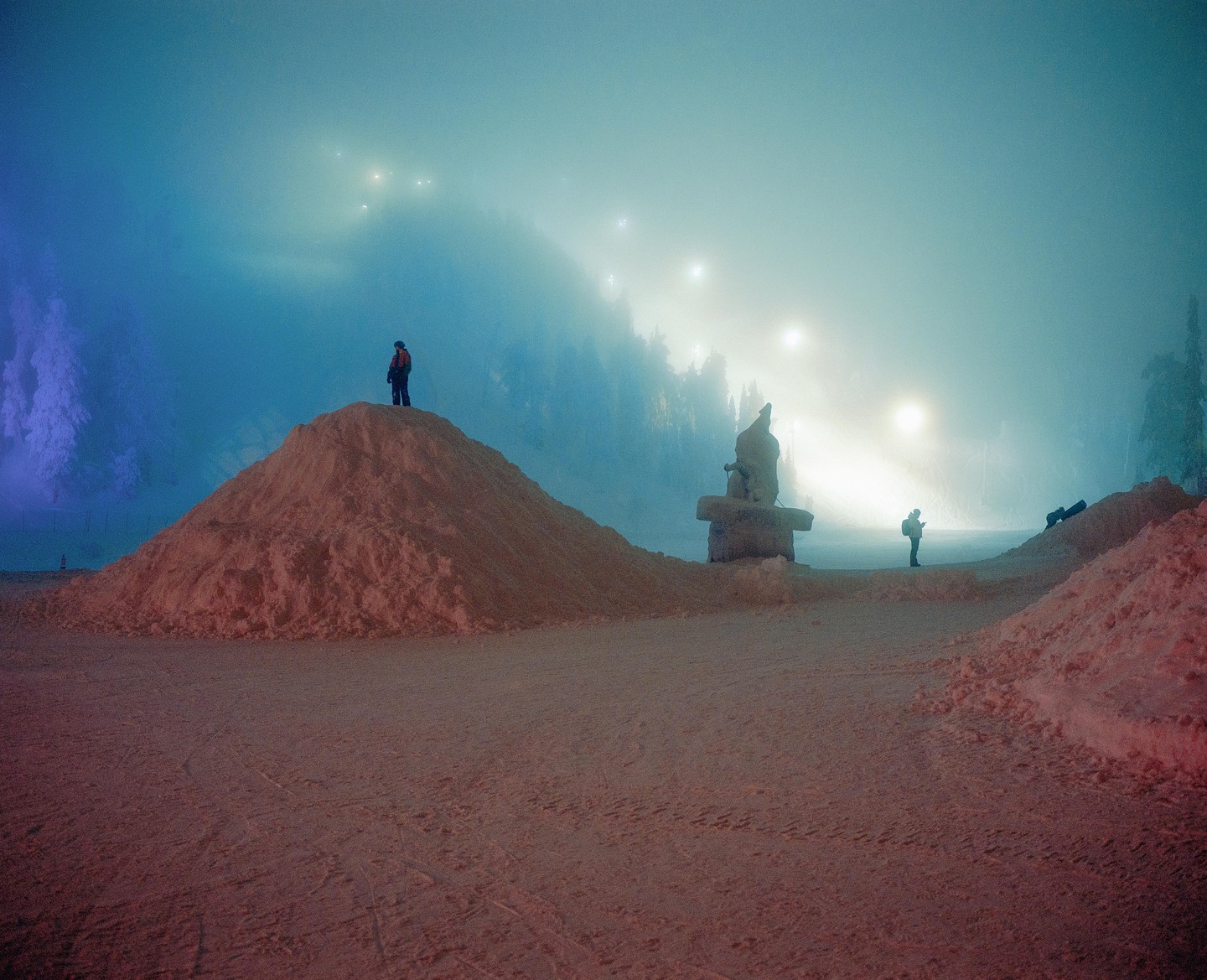 A person stands atop a snow hill, looking out at a foggy, snow-covered landscape with distant lights.