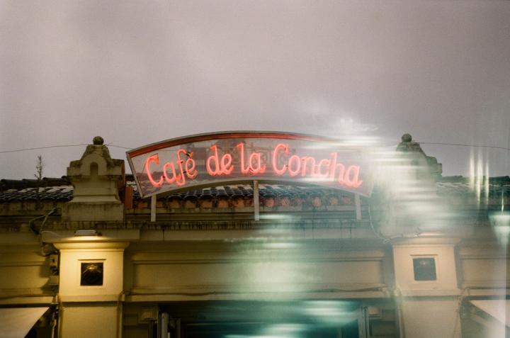A neon sign for "Cafe de la Concha" is mounted on a building under a cloudy sky.