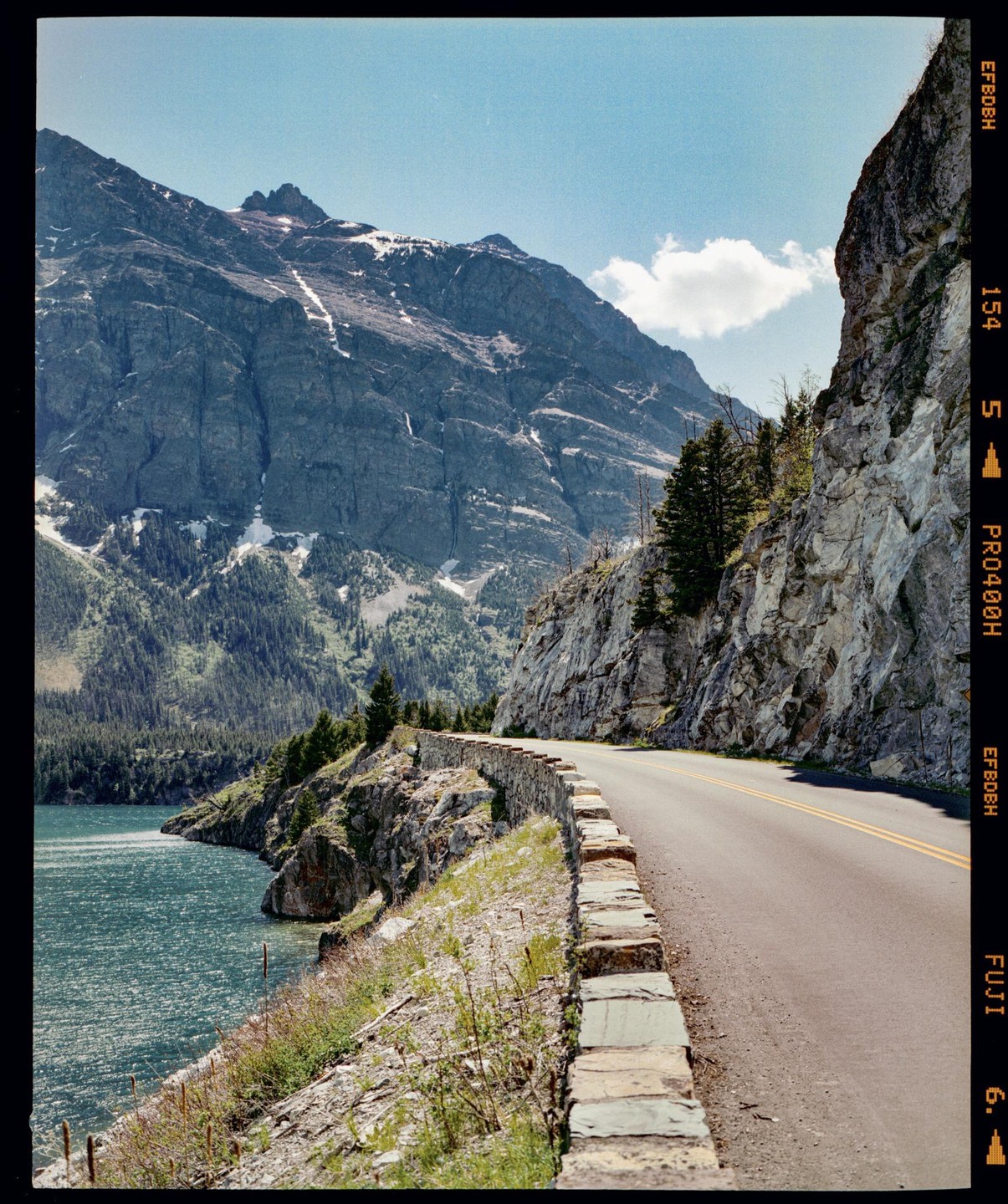 A winding road with a stone wall runs alongside a lake with mountains in the background.
