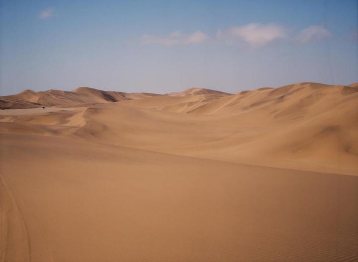 A vast desert landscape features rolling sand dunes under a clear sky with a few clouds.