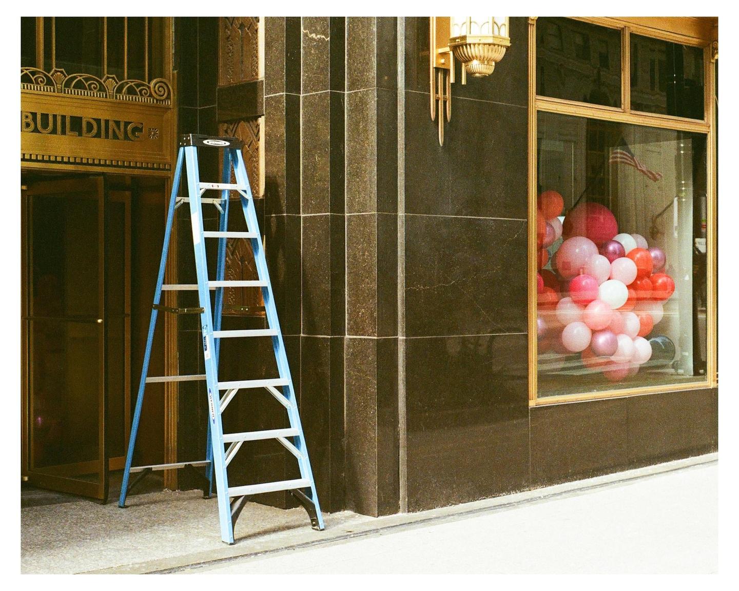 A blue ladder stands in front of a building entrance next to a window display filled with balloons.