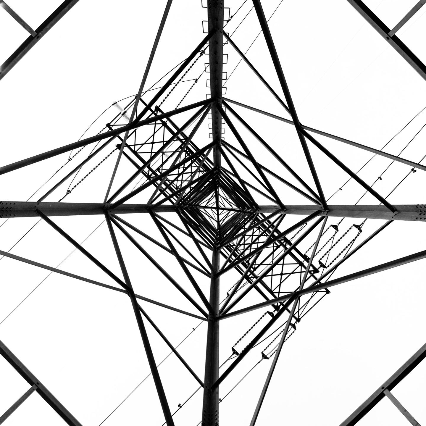 A low-angle view of a power line tower shows its symmetrical structure against a white sky.