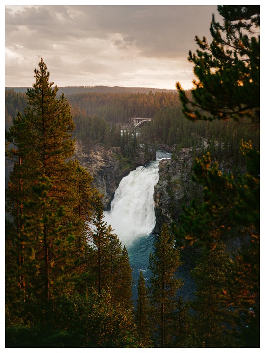 A waterfall cascades into a river surrounded by a forest with a bridge in the distance under a cloudy sky.