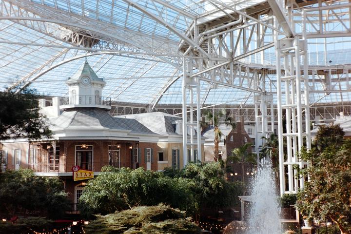 A large indoor atrium with a glass roof features a clock tower building, lush greenery, and a fountain.