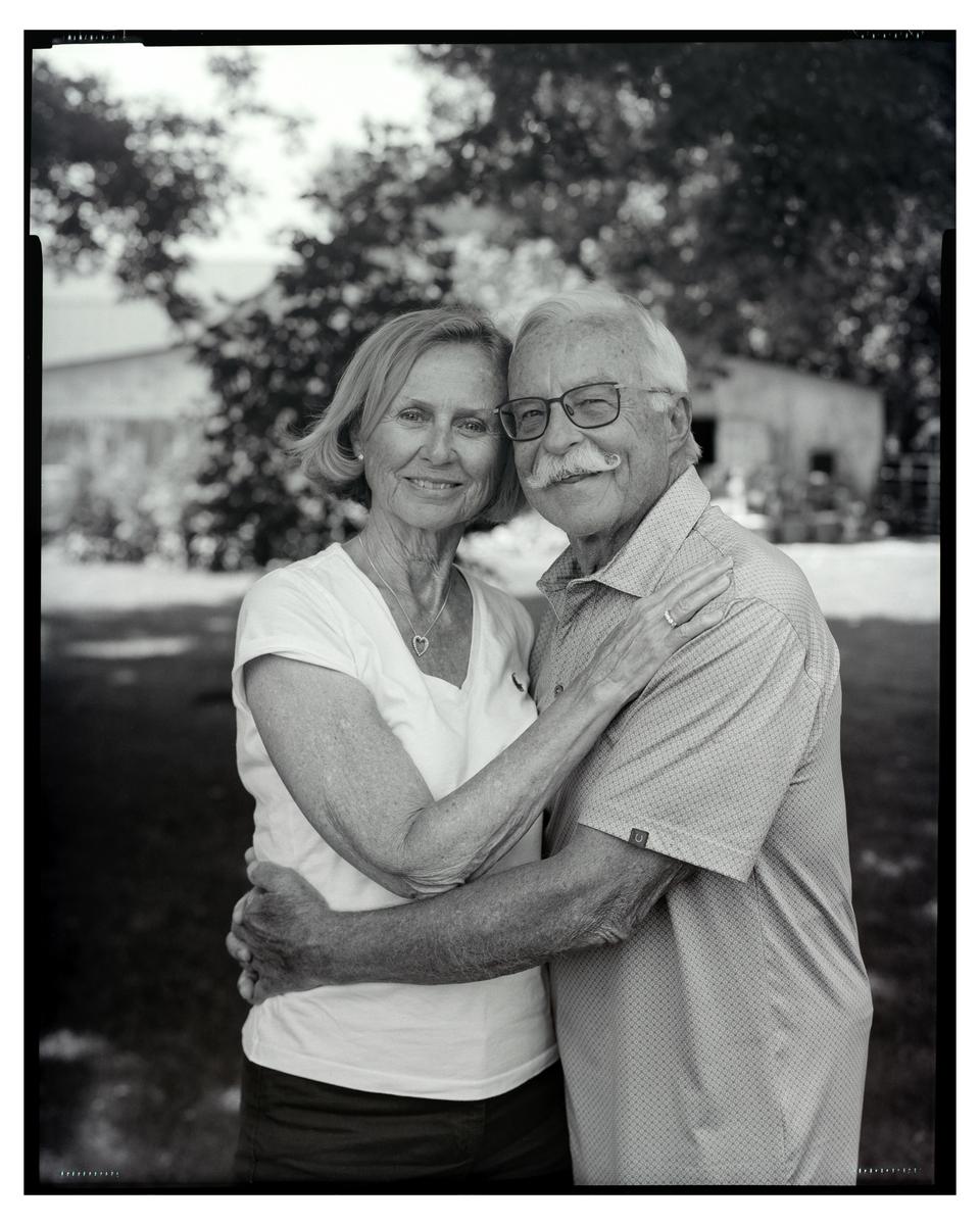 A man and woman embrace and smile at the viewer in a black and white outdoor portrait.