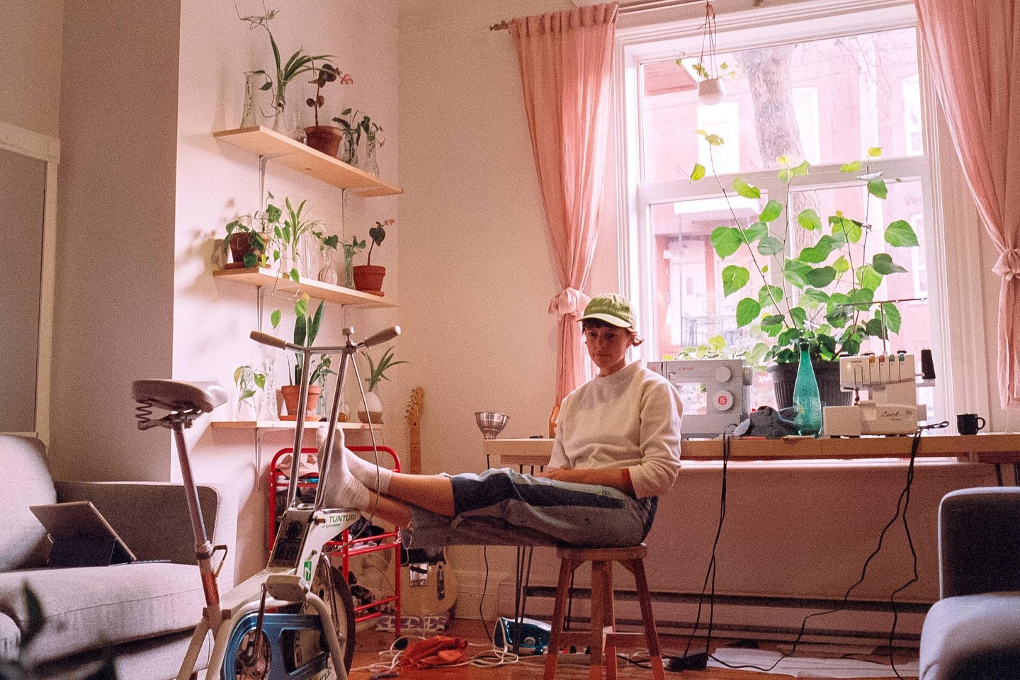 A woman sits on a stool with her feet resting on an exercise bike in a sunlit room filled with plants.