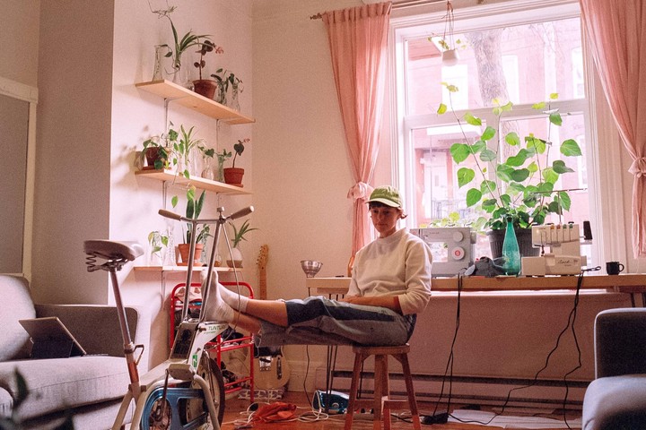 A woman sits on a stool with her feet resting on an exercise bike in a sunlit room filled with plants.