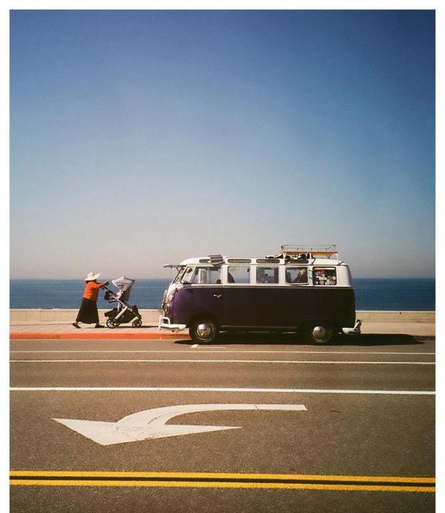 A woman pushes a stroller past a purple and white van parked next to a road with an ocean in the background.