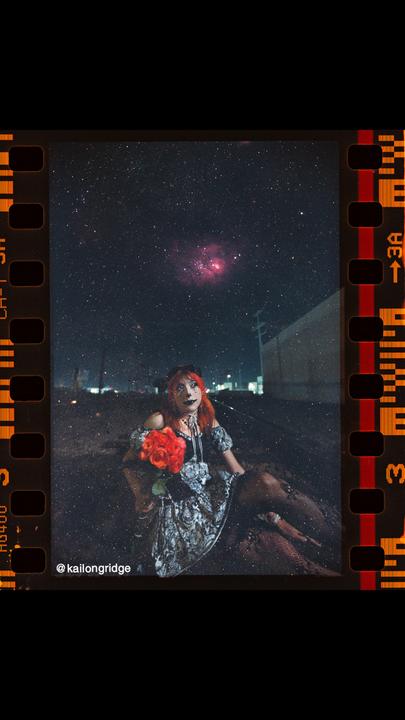 A woman with red hair and dark makeup holds a bouquet of red roses while sitting on the ground under a starry night sky.