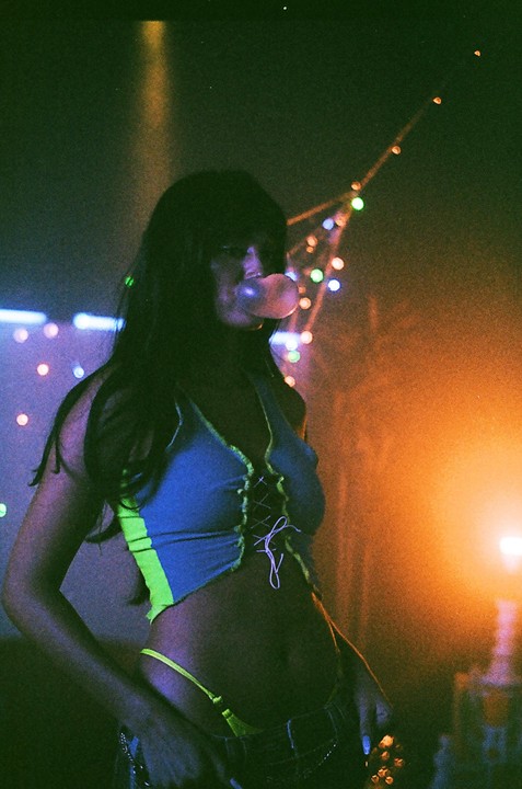 A woman blows a bubble gum bubble in a dimly lit room with colorful string lights.