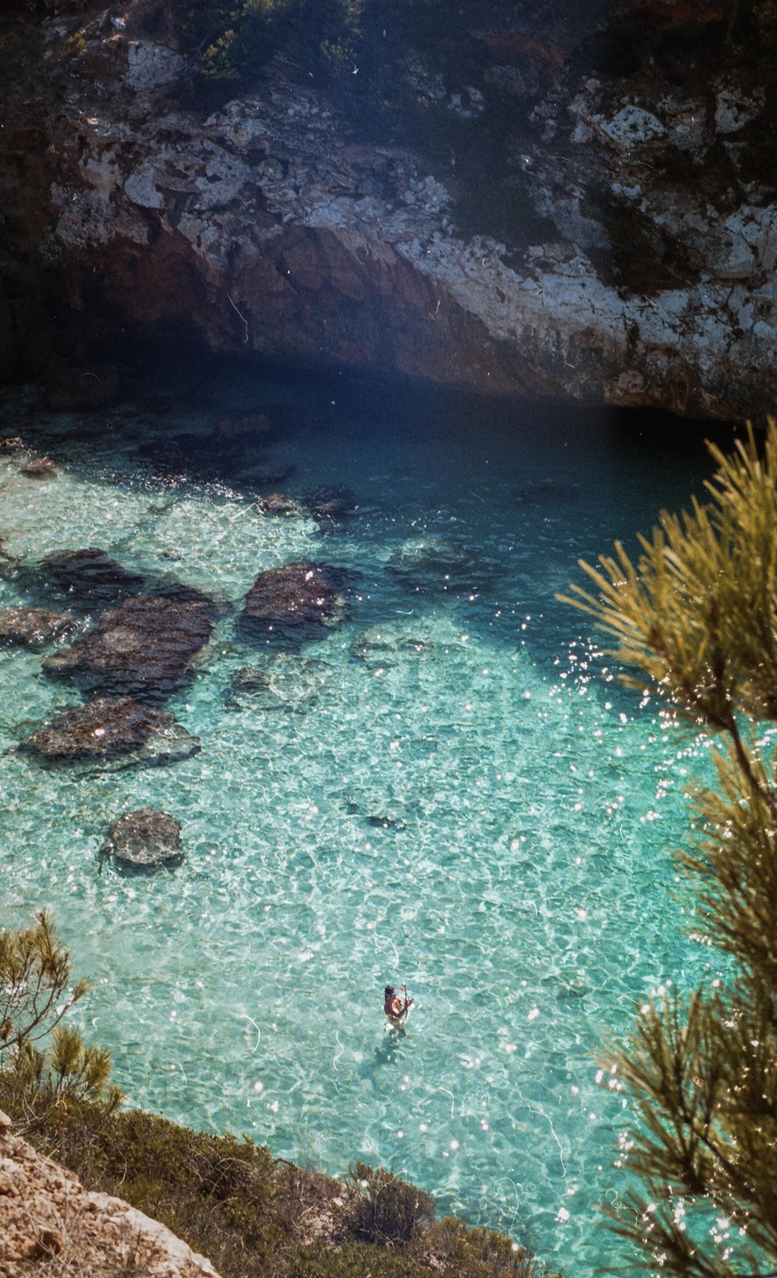 A person stands in clear turquoise water surrounded by rocky cliffs and green foliage.
