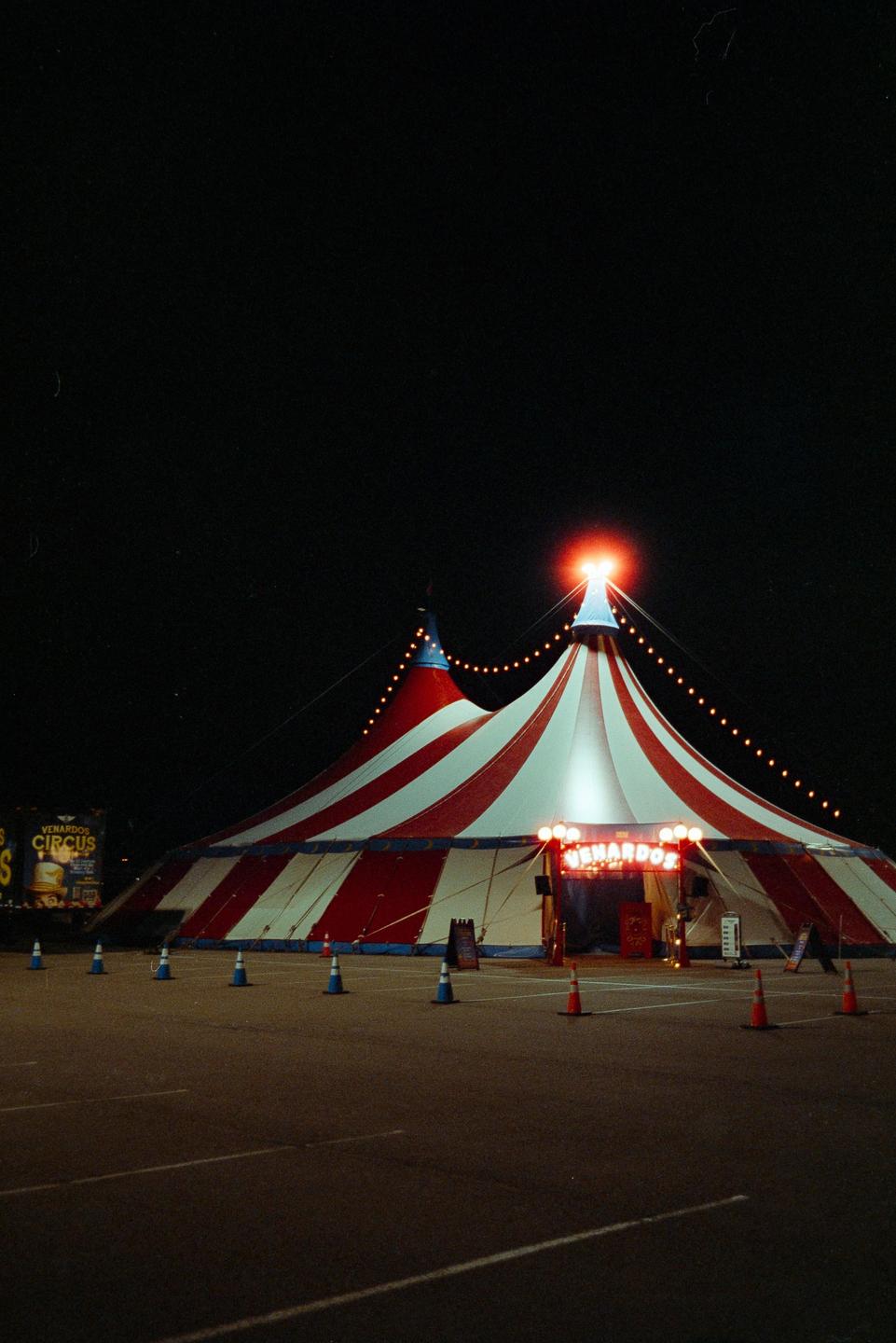 A red and white striped circus tent is illuminated at night, with a sign for "Venardos Circus" visible on a truck in the background.