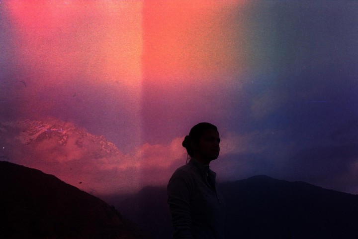 A woman stands silhouetted against a vibrant, colorful sky with mountains in the background.