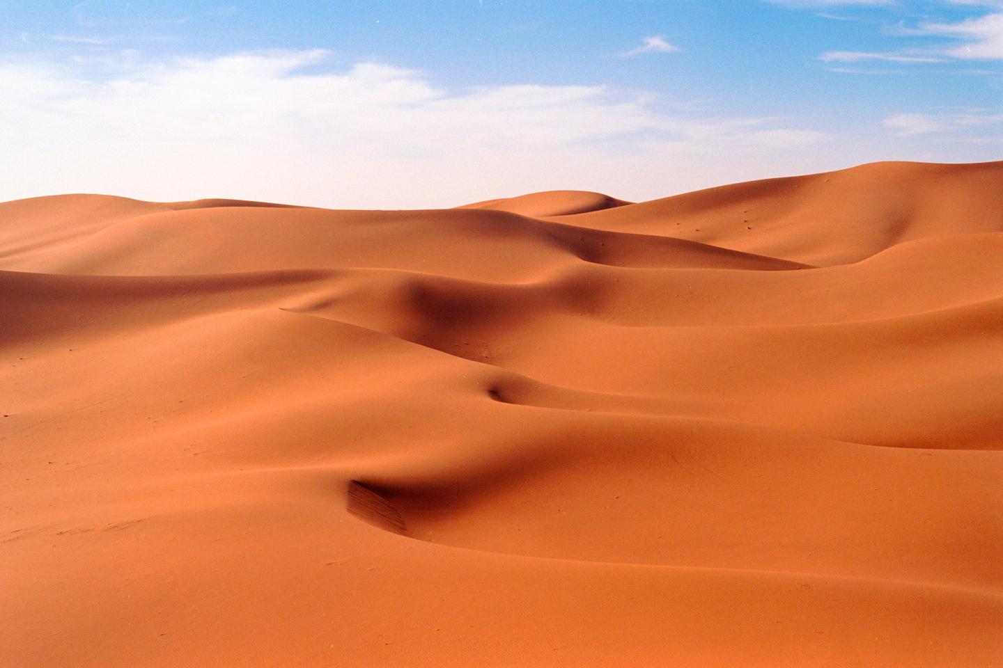 Rolling sand dunes stretch across the desert under a blue sky with scattered clouds.