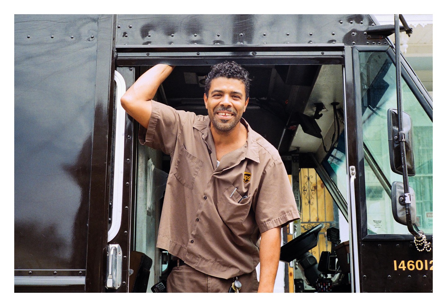 A man in a brown uniform smiles while leaning out of a delivery truck.