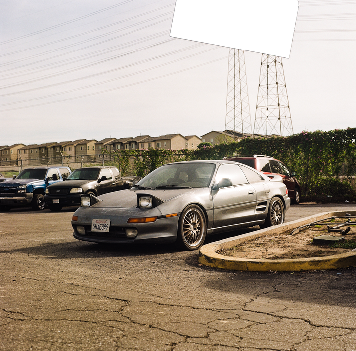 A gray sports car with pop-up headlights is parked on cracked asphalt next to a yellow curb.