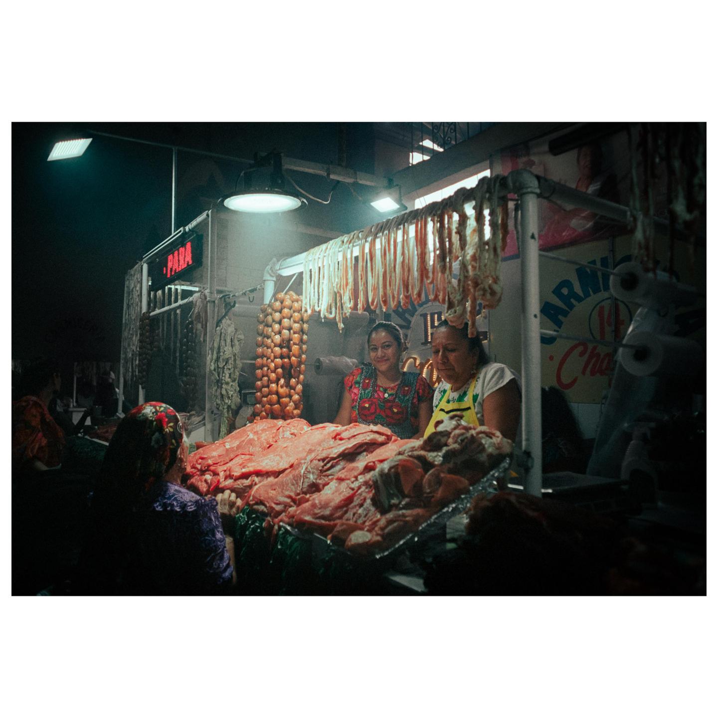 Two women stand behind a counter laden with meat and sausages in a market stall.