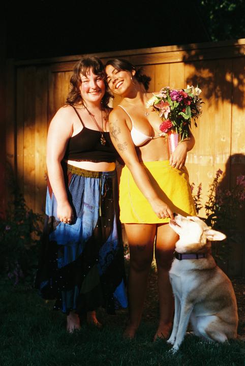 Two women stand in a sunlit yard with a dog and a bouquet of flowers.