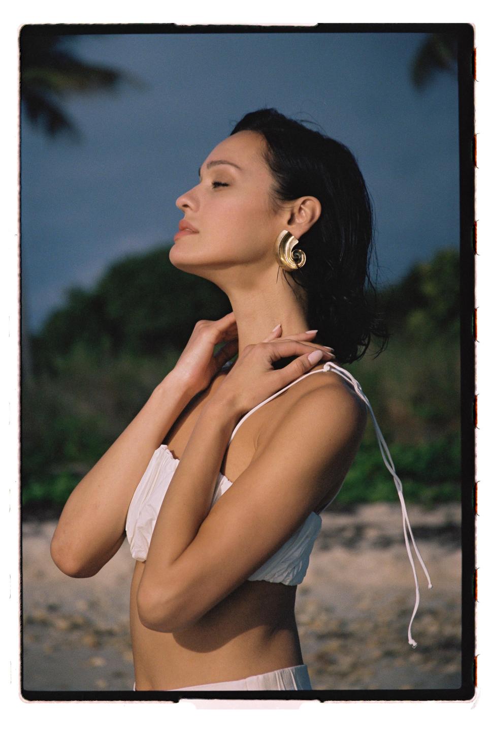 A woman with her eyes closed and head tilted back stands on a beach with trees in the background.