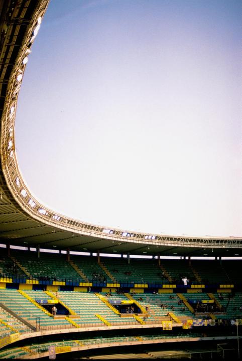 A large, empty stadium with green and yellow seats is visible under a bright sky.