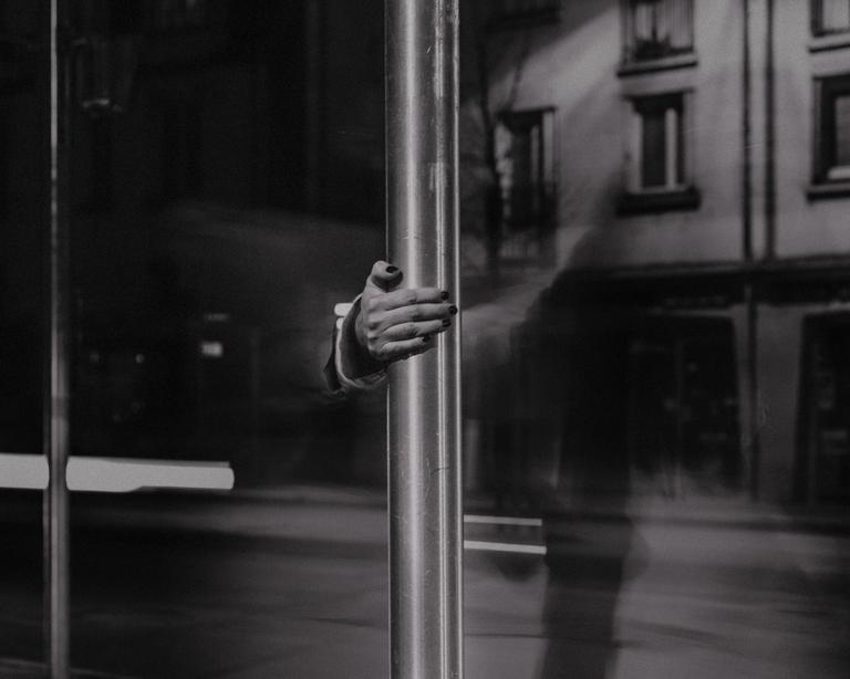 A hand with dark nail polish holds a metal pole in front of a reflective surface with building reflections.